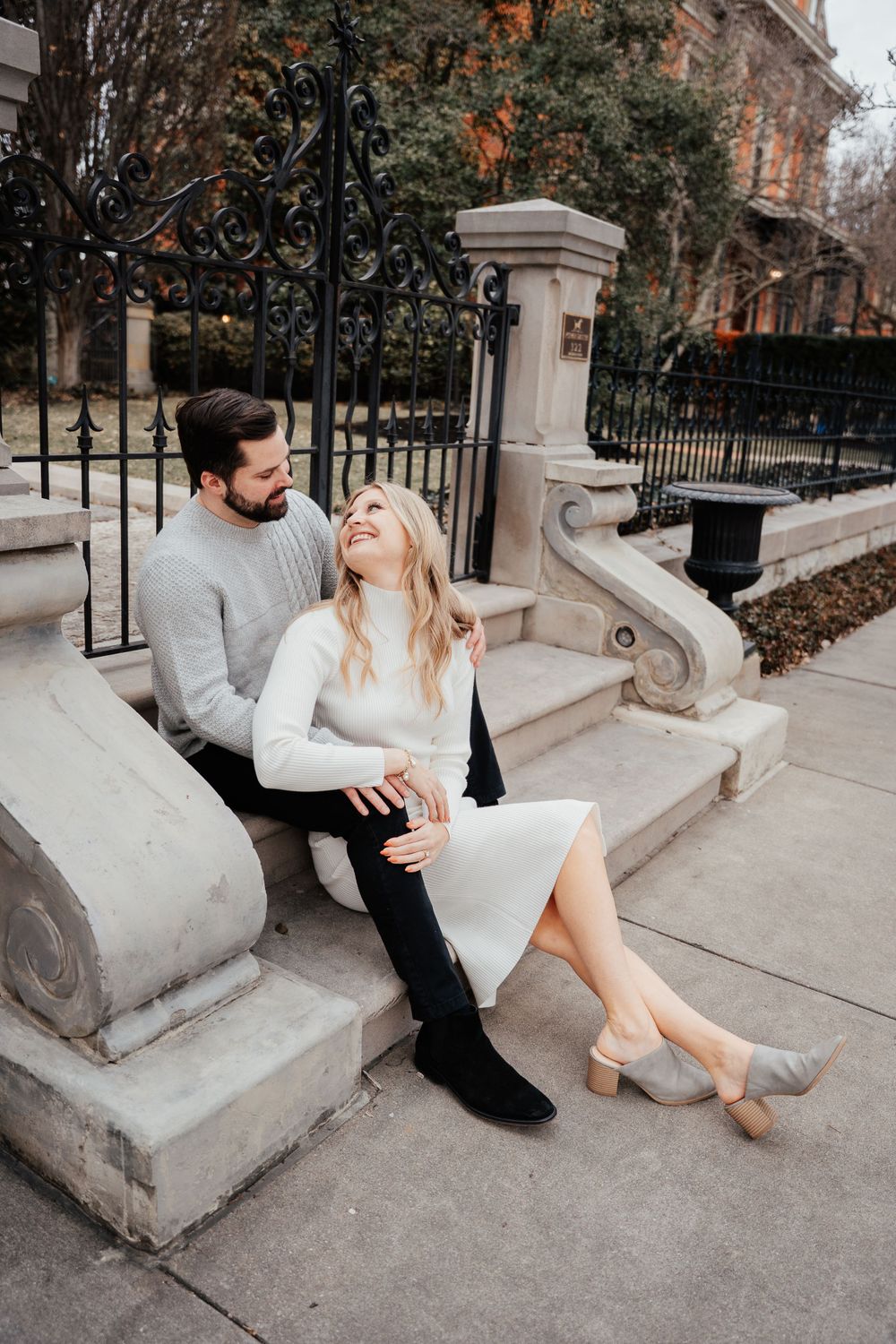 A couple sits together on stone steps outside an ornate building with wrought iron fencing during an autumn day.