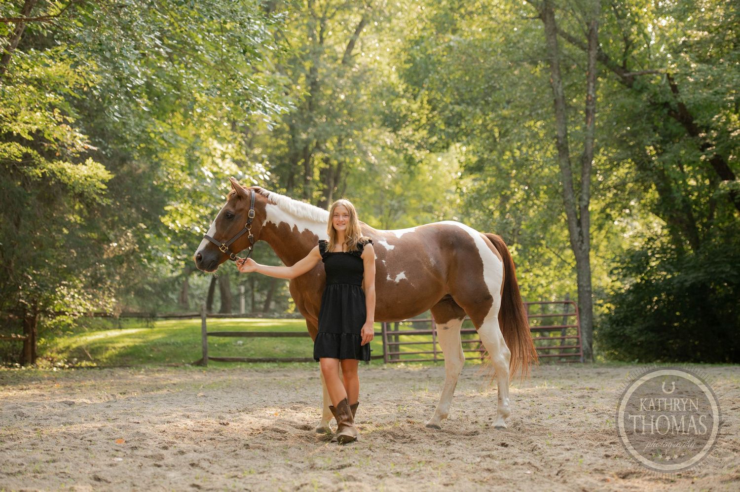 A paint horse and person in a black dress stand together in a sunlit wooded area.