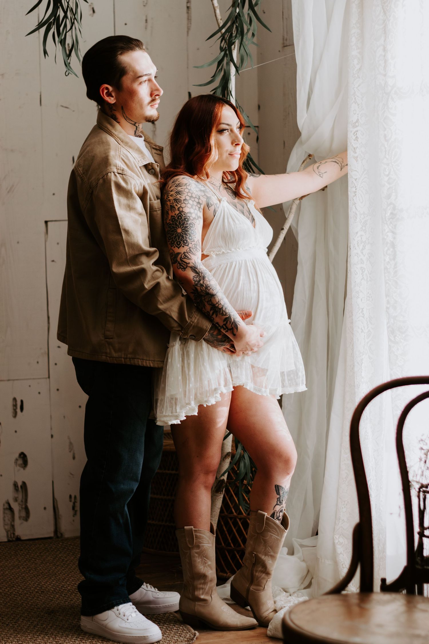 A couple stands by a window, with someone wearing a white lace dress and brown boots while expecting a baby.