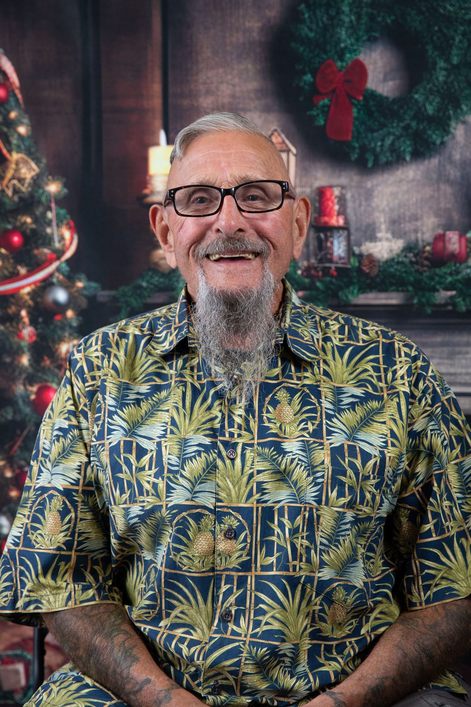 A person wearing glasses and a tropical print Hawaiian shirt poses in front of Christmas decorations and a wreath.
