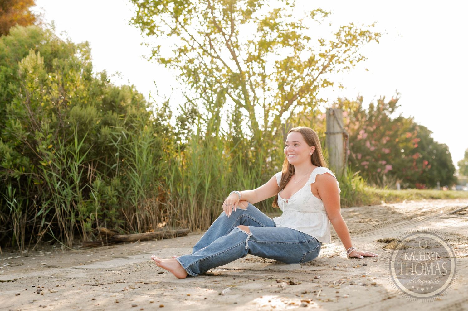 A casual outdoor portrait sitting on a sandy path with green foliage and warm sunlight.