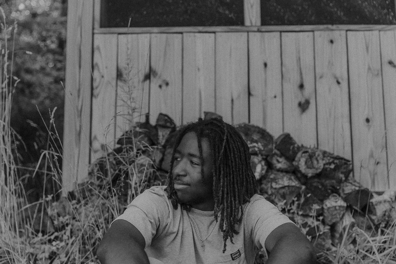 A person with dreadlocks sits outdoors against a wooden fence in a black and white photograph.