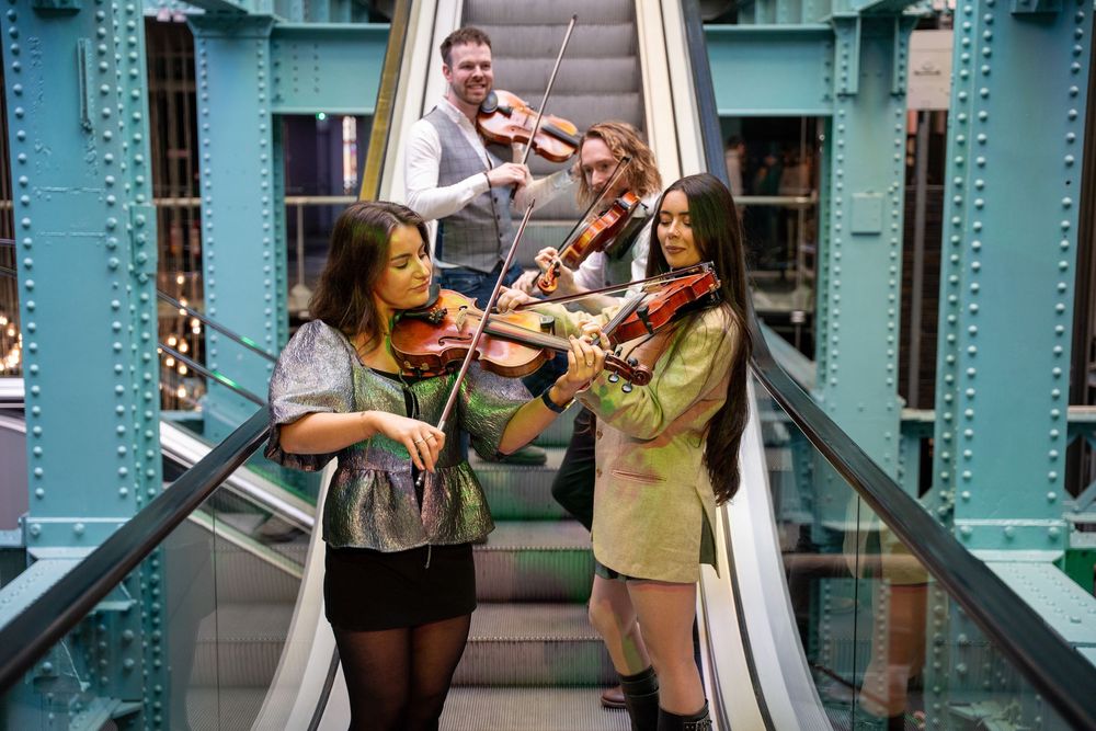 Musicians perform with violins on an escalator surrounded by aqua-colored architectural details.