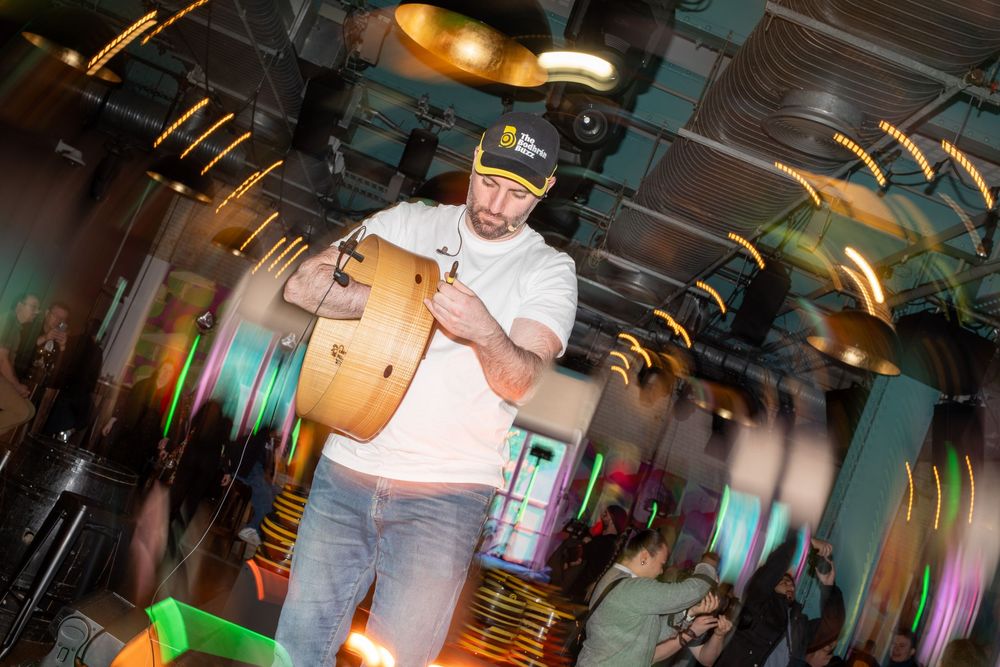A musician performs with a drum in a dimly lit venue with colorful light trails in the background.