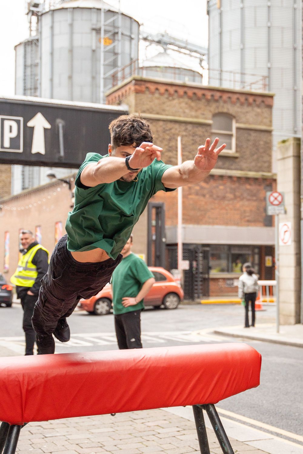 A person in green performs a parkour leap over a red street barrier in an urban setting.