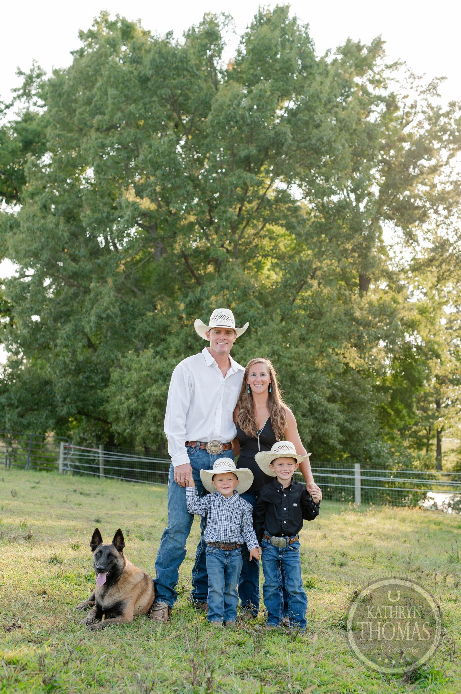 Family of four poses with German Shepherd dog in front of large tree during golden hour.