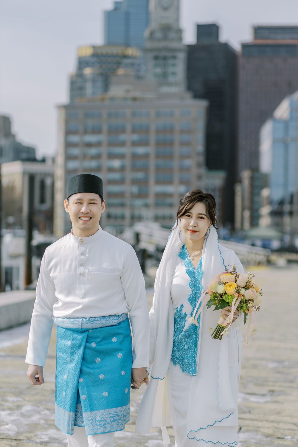 Traditional wedding couple in white and turquoise attire photographed against city skyline.