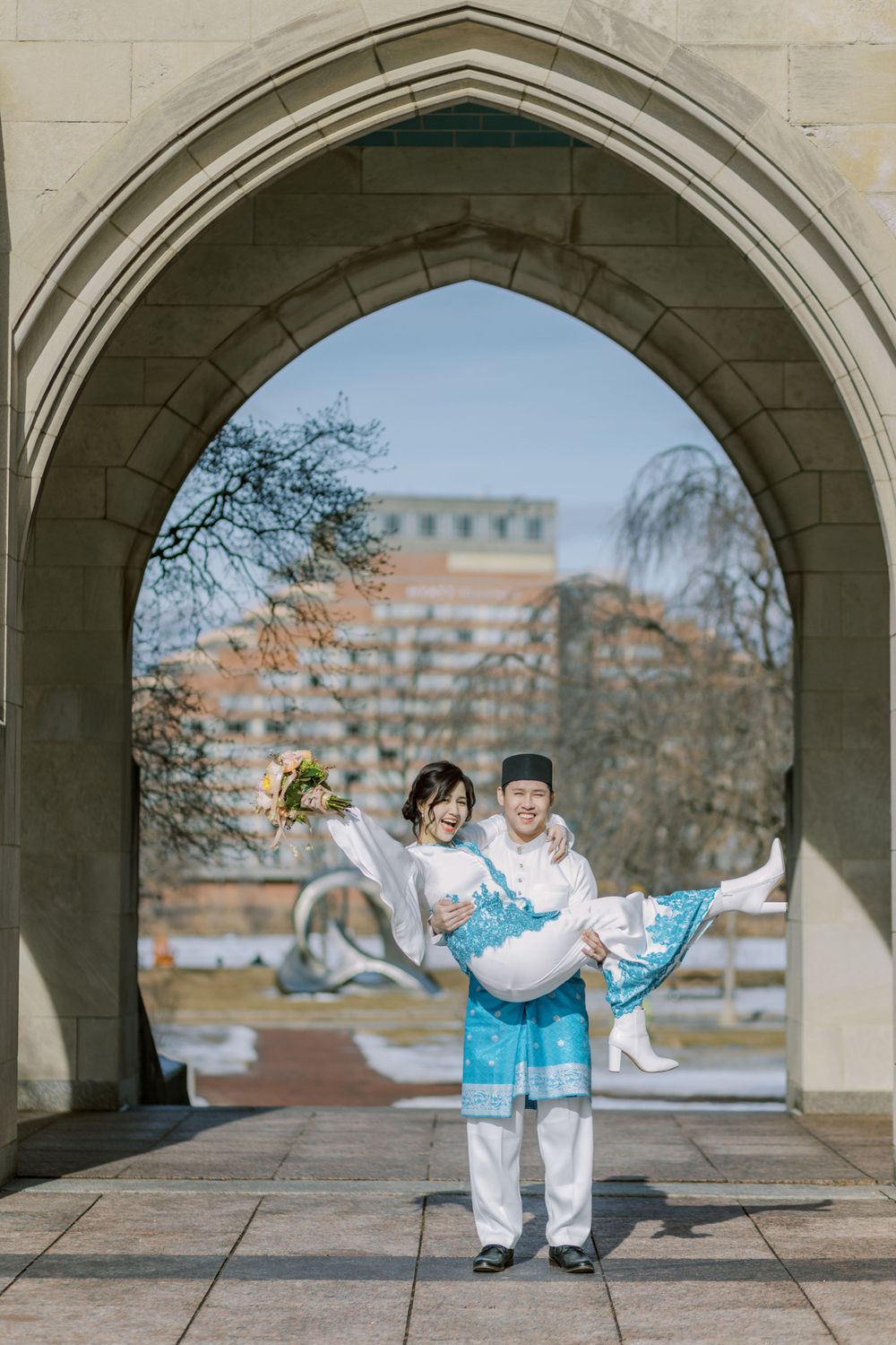Romantic couple poses playfully under stone archways in matching white and blue traditional outfits.