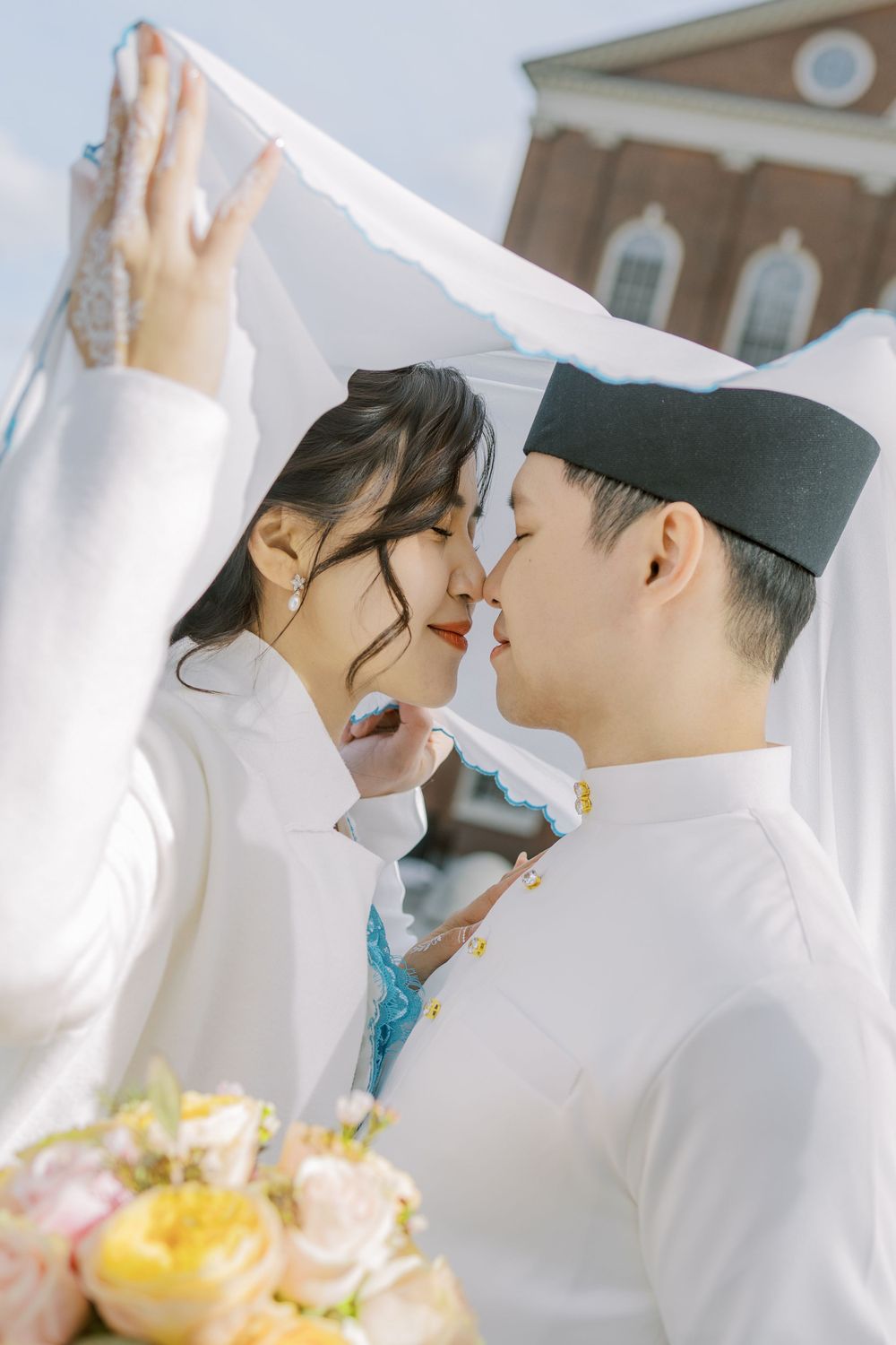 Intimate couple portrait with traditional white outfits and black cap under flowing white fabric.
