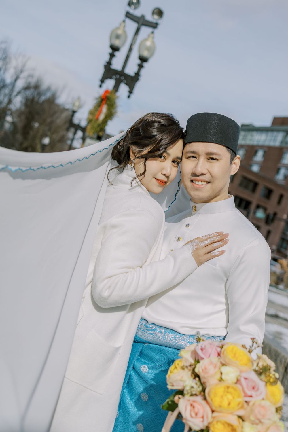Couple in traditional Korean-inspired attire pose with colorful bouquet against urban backdrop.