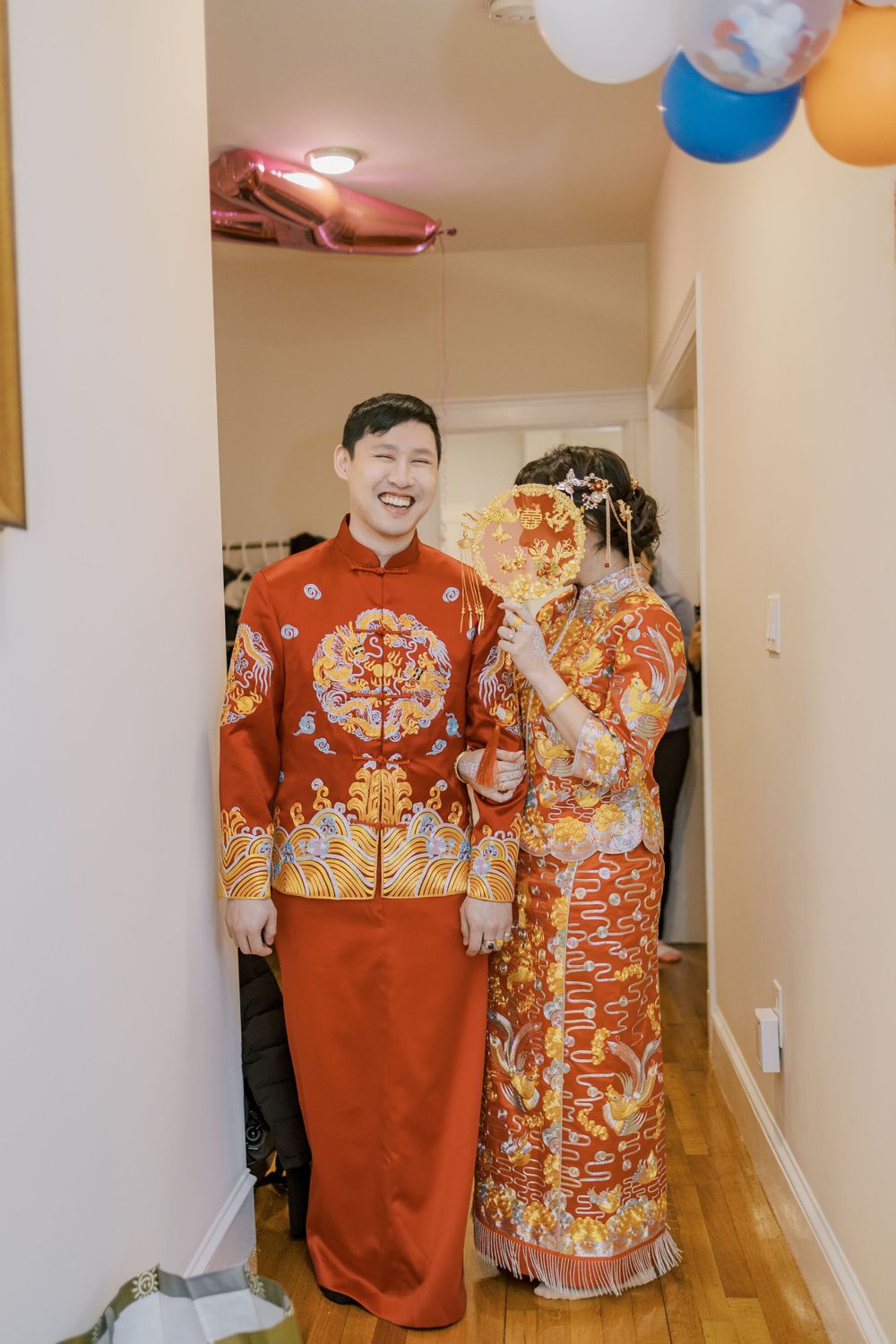 Couple in traditional Chinese wedding attire laughing together in decorated hallway with balloons.