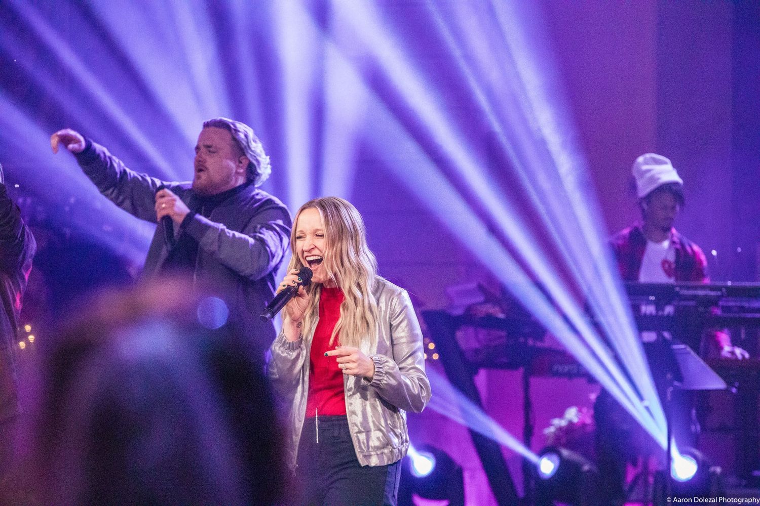 Vocalist performs on stage in silver jacket with dramatic purple lighting and band members in background.