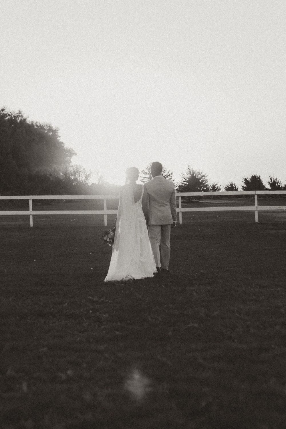 A couple in wedding attire walks together across a grassy field at sunset with white fencing in the background.