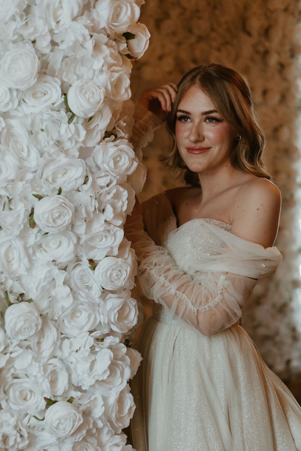 An elegant bridal portrait against a backdrop of white roses featuring a romantic off-shoulder gown.