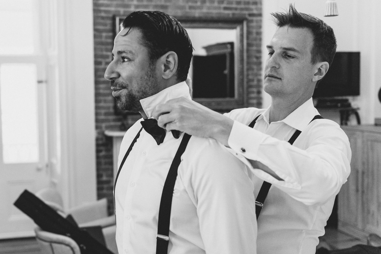 A groomsman in a white dress shirt helps adjust a bow tie and suspenders in a black and white photograph.