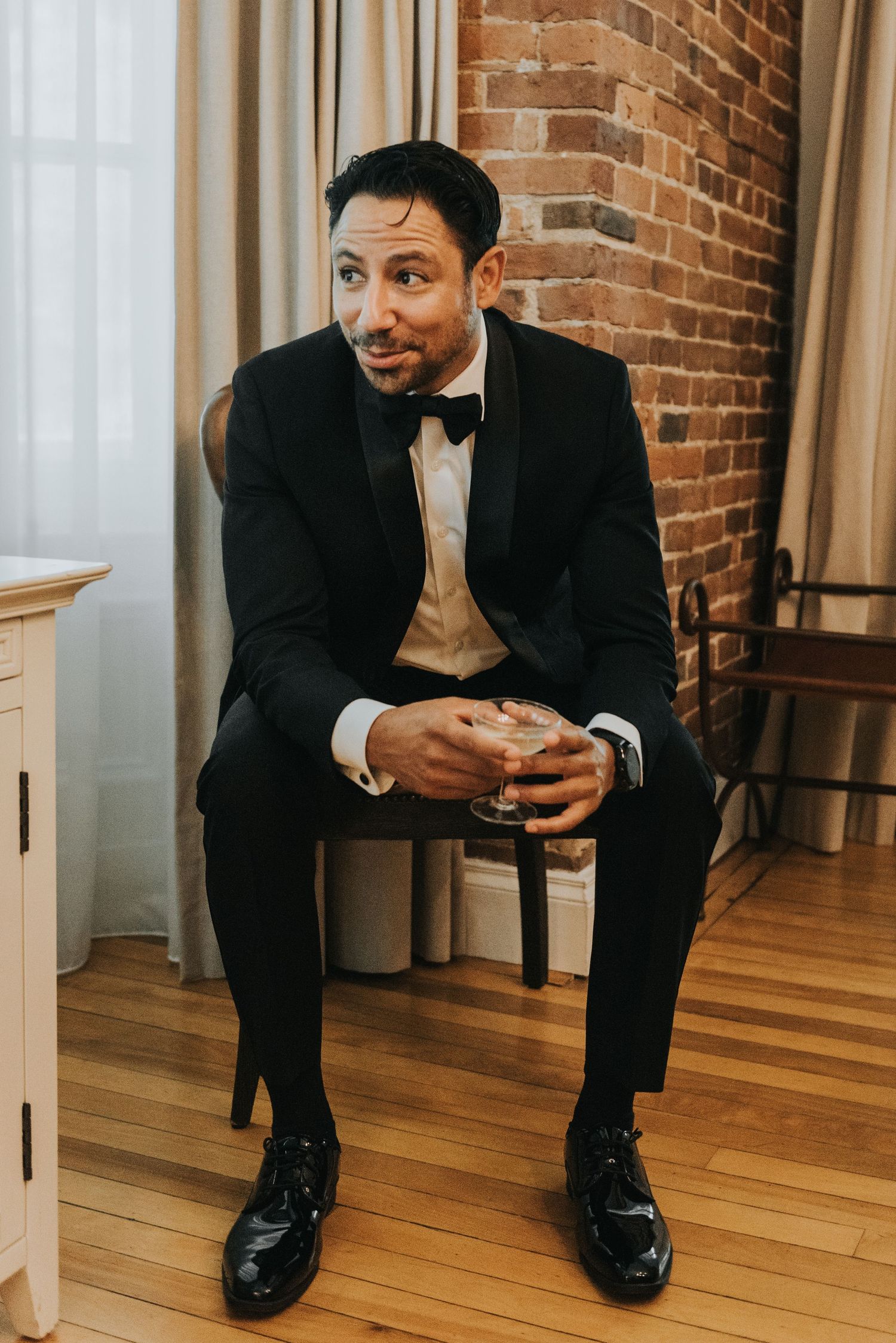 A person in a black tuxedo and bowtie sits on a chair in an elegant room with exposed brick walls and hardwood floors.