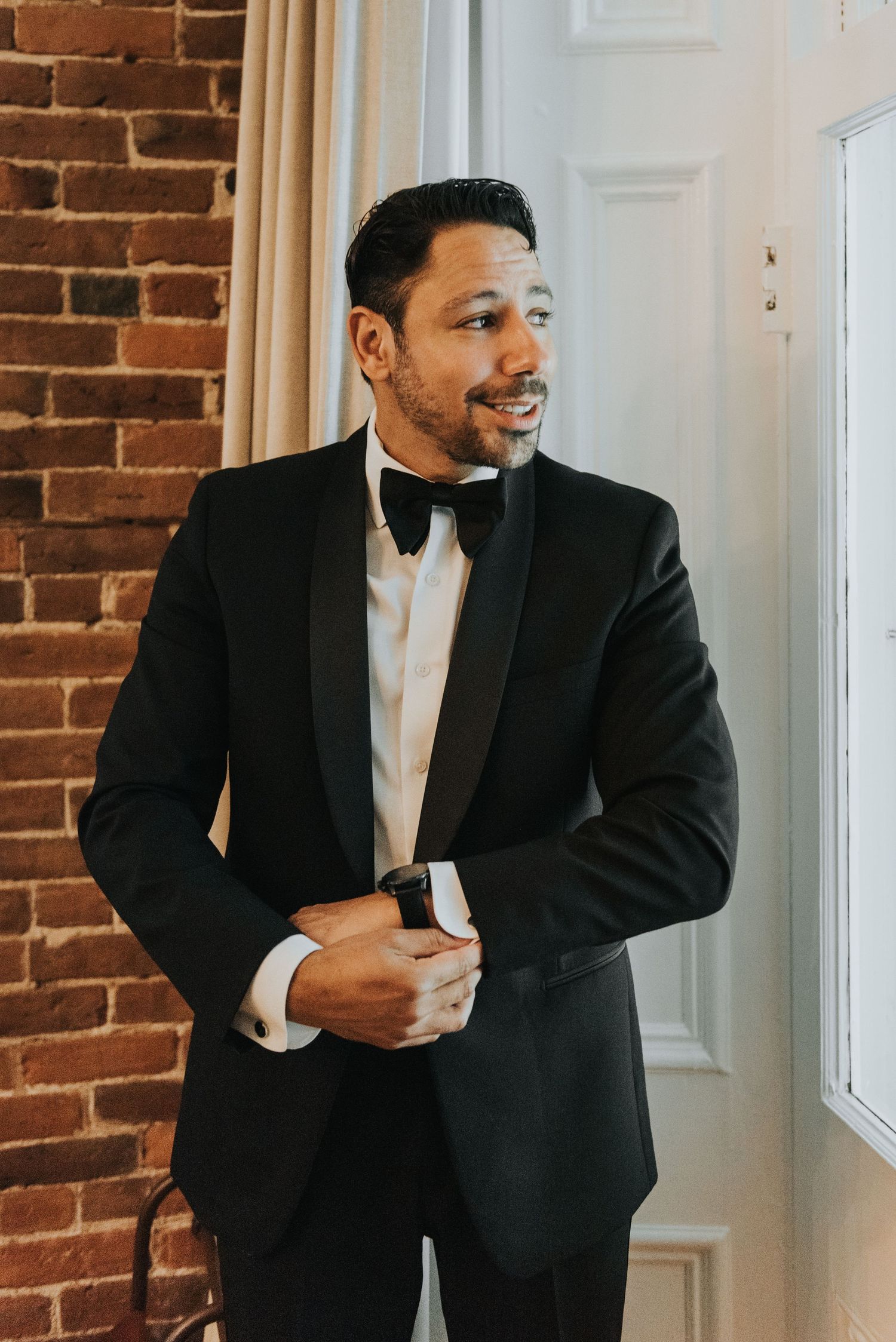 Elegant groom in classic black tuxedo with bow tie stands against exposed brick wall near window in formal setting.
