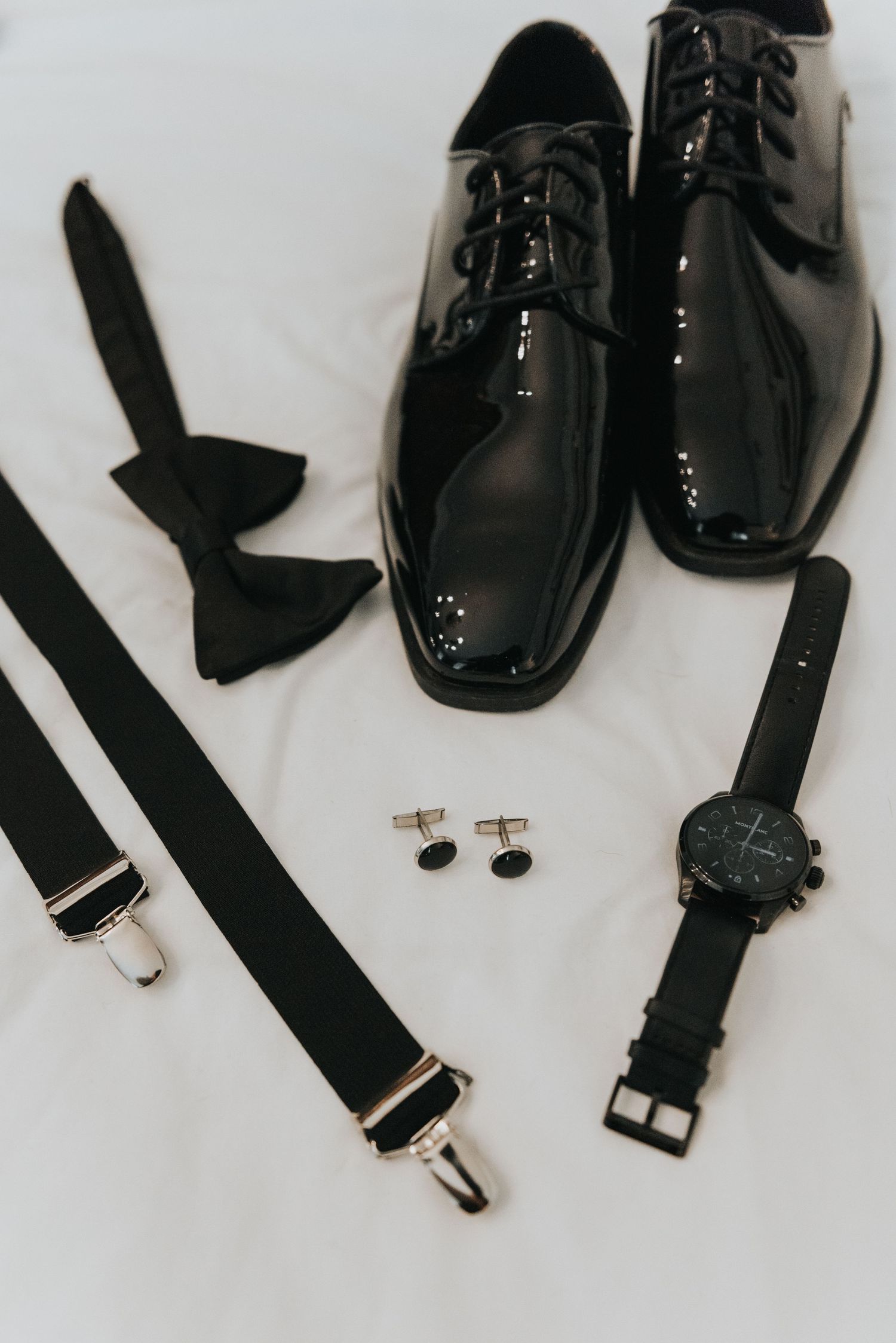 Formal black accessories including patent leather dress shoes, bow tie, suspenders, watch and cufflinks arranged on white surface.