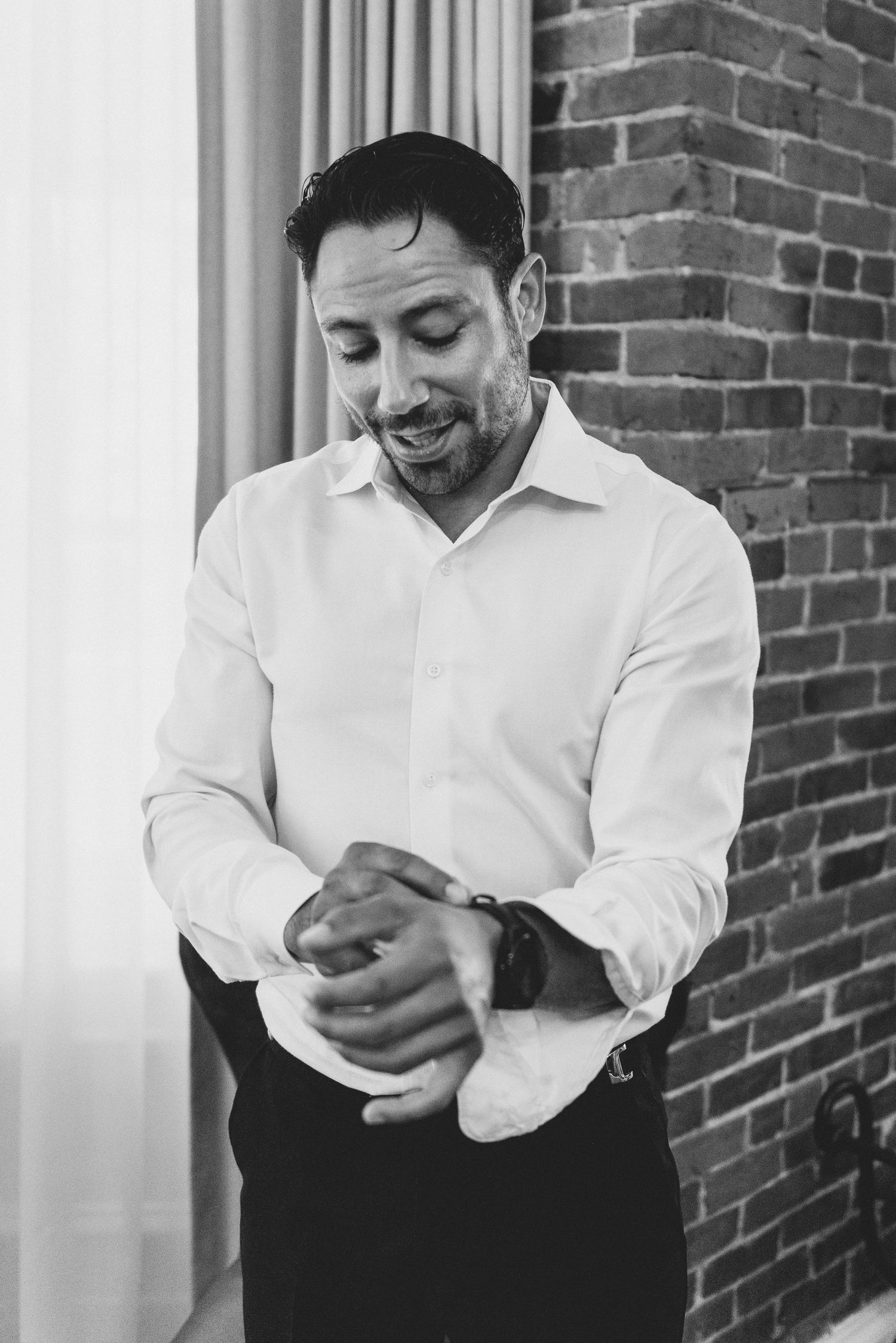 A person in a white dress shirt adjusts their cuff while smiling and standing against a brick wall background.