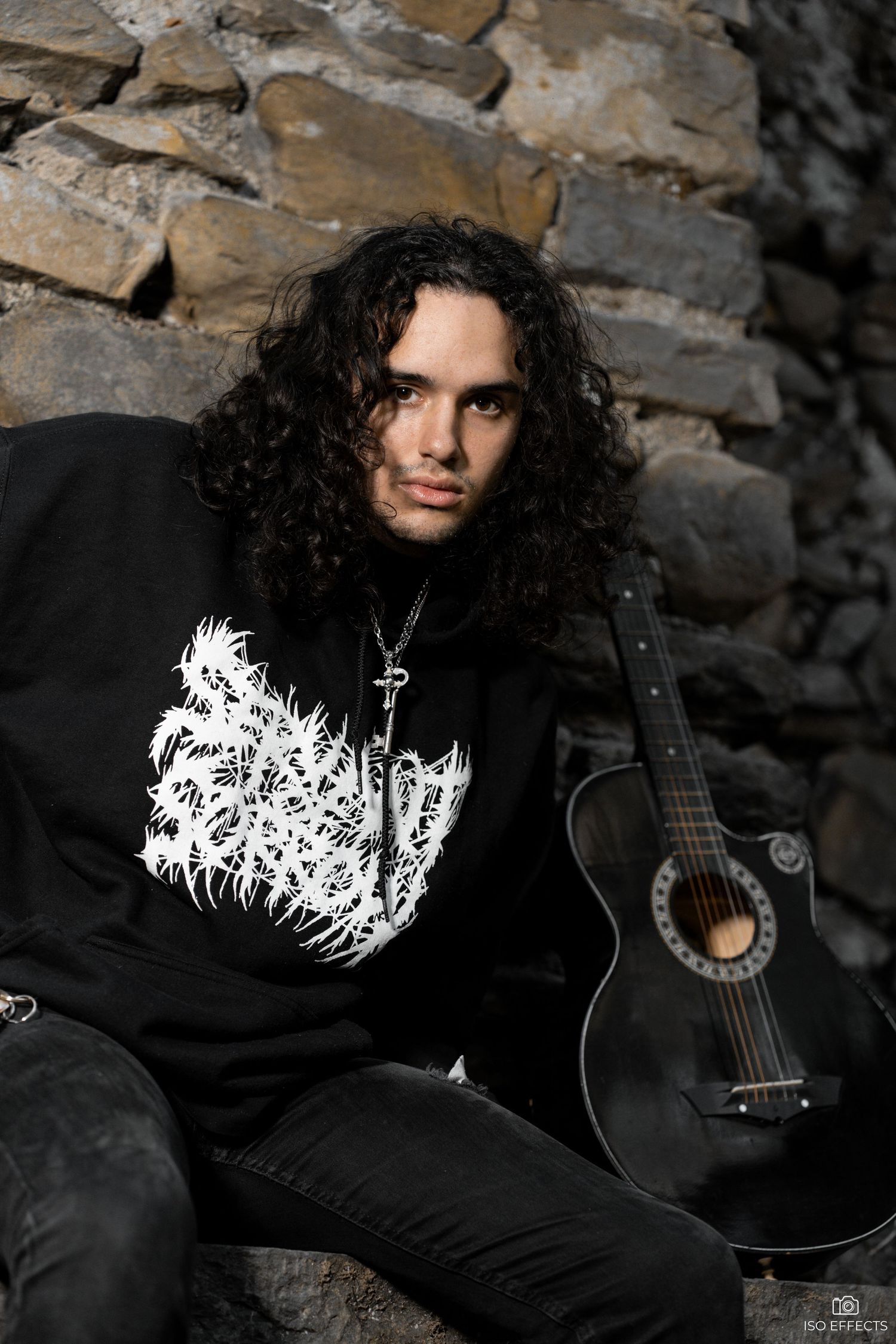 Musician in black heavy metal shirt poses with acoustic guitar against stone wall backdrop.