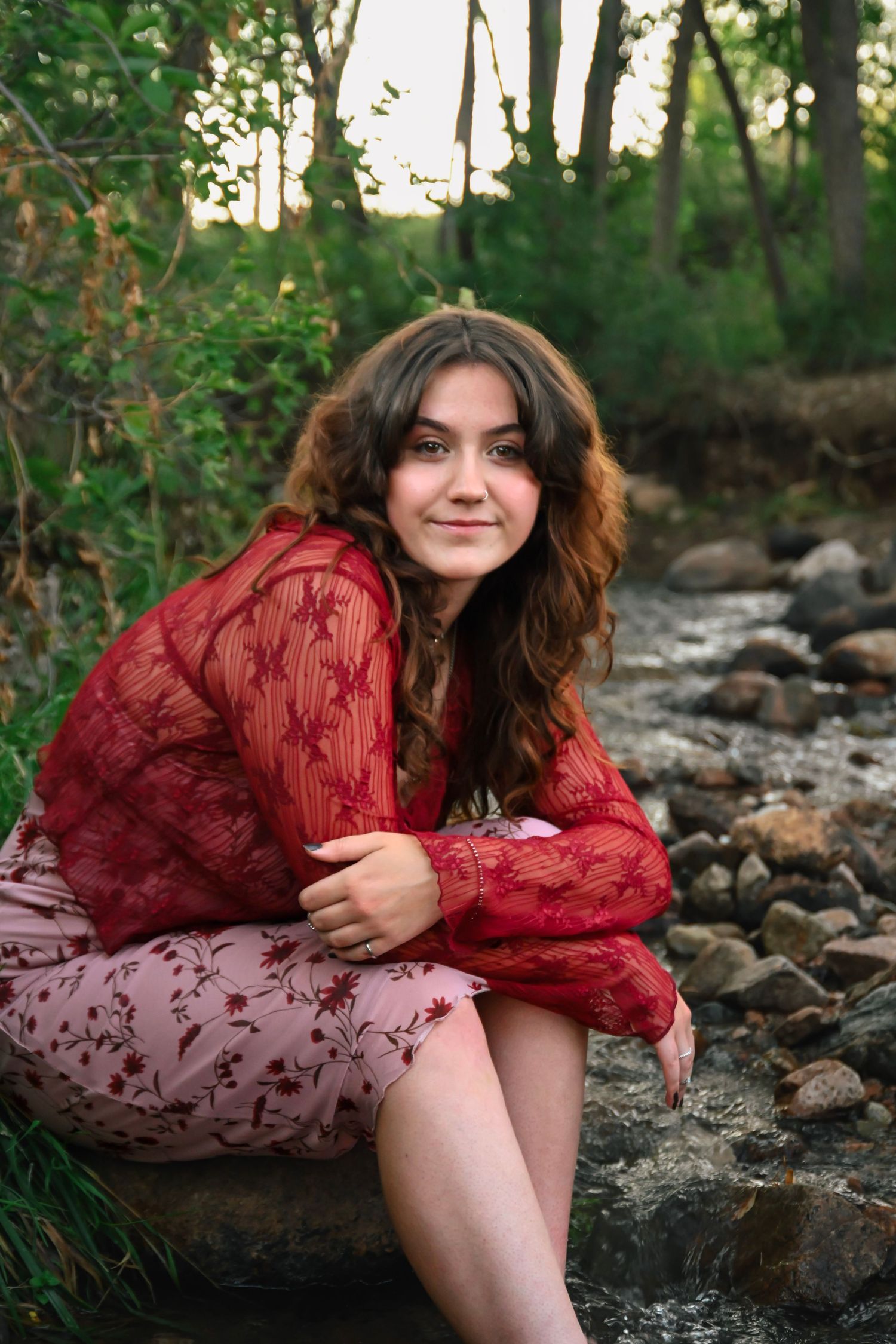 Young woman in red lace top and floral skirt sitting on rocks by a forest stream, smiling.