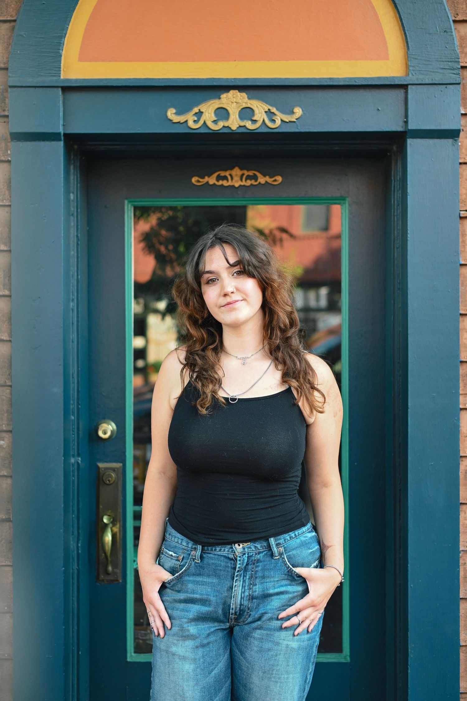 Young woman with curly hair in black tank top and jeans posing in front of decorative teal door.