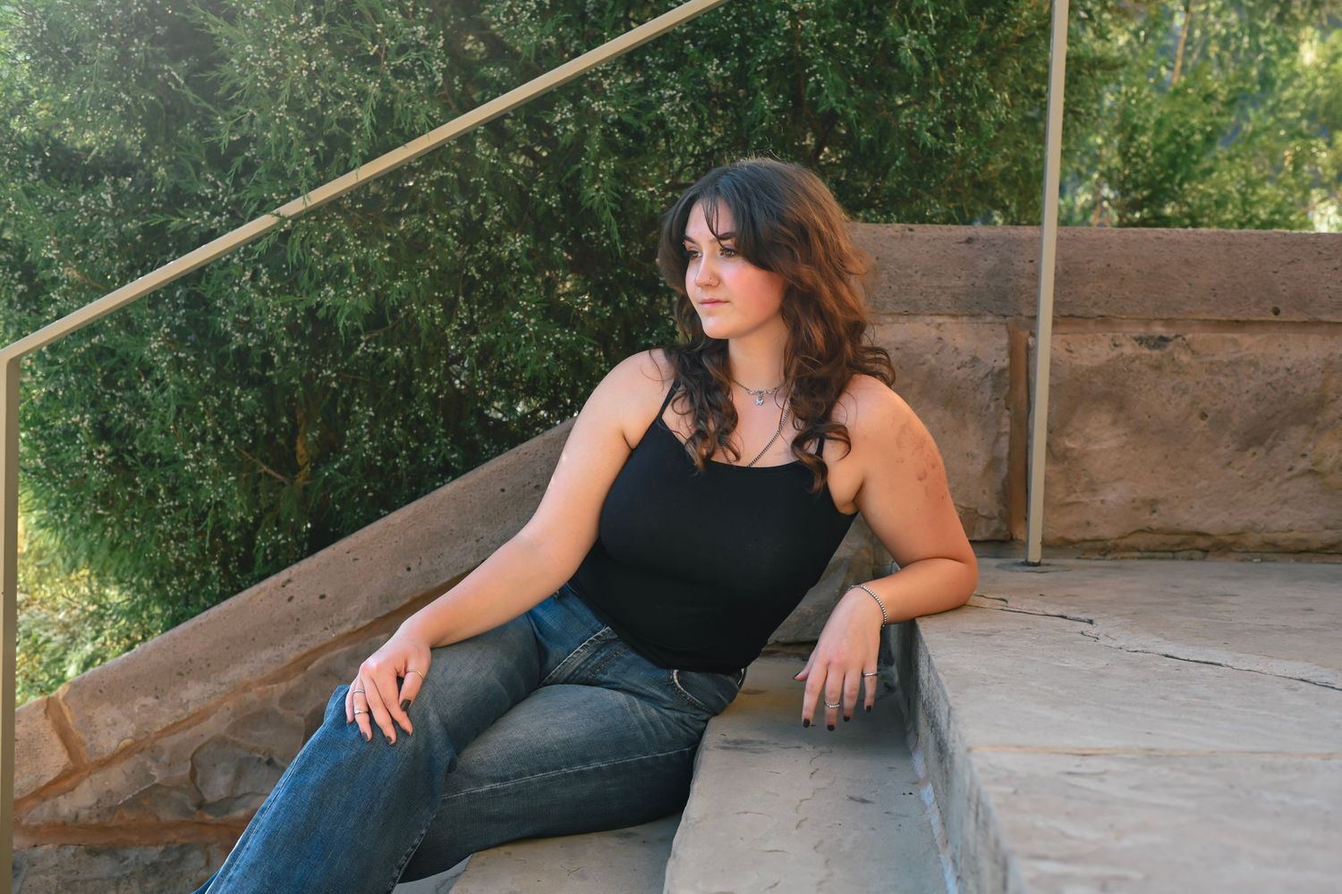 Young woman in black tank top and jeans sitting on stone steps outdoors with lush green trees behind her.