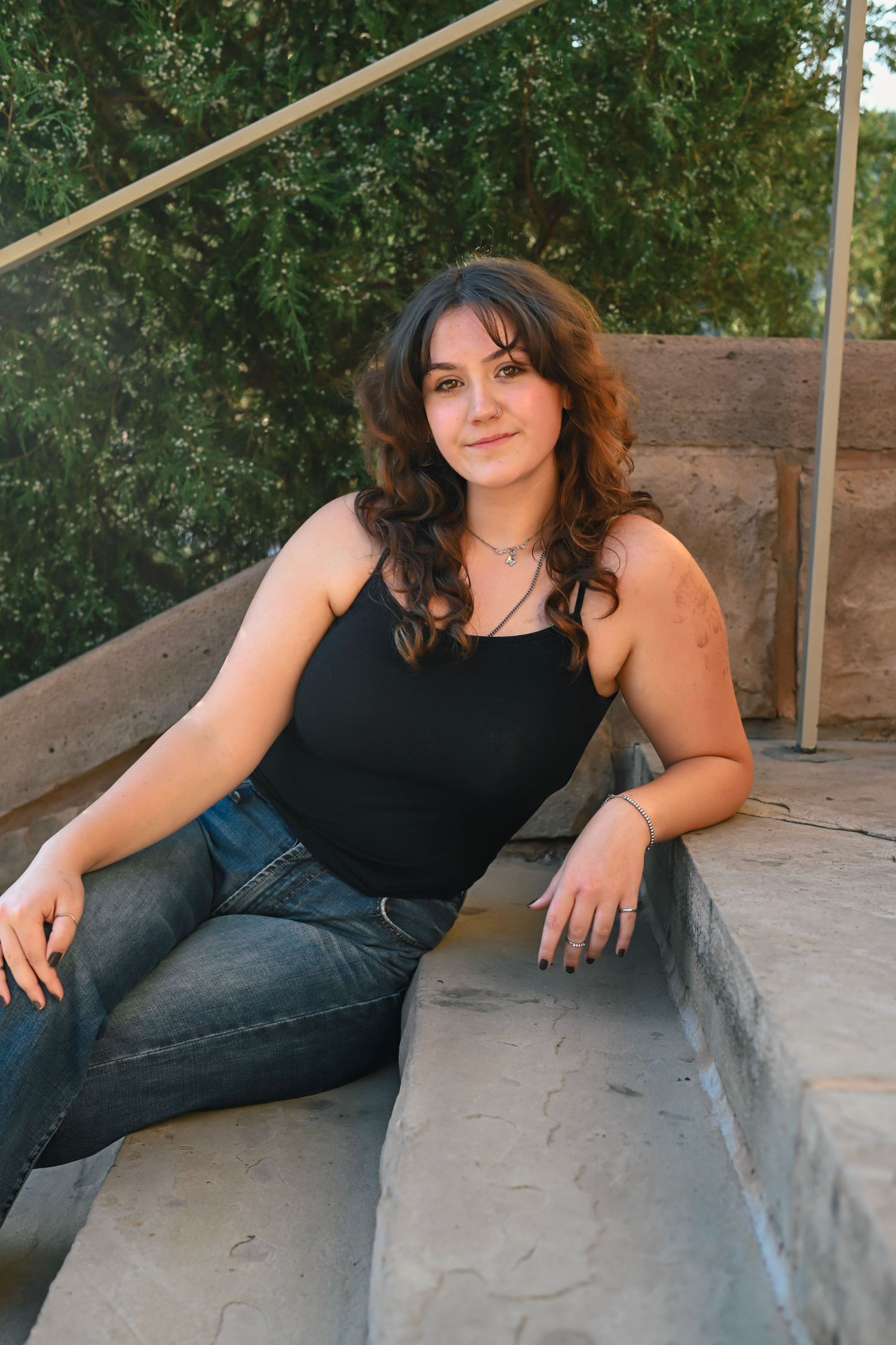 Young woman with curly brown hair sitting on stone steps outdoors, wearing a black tank top and jeans.