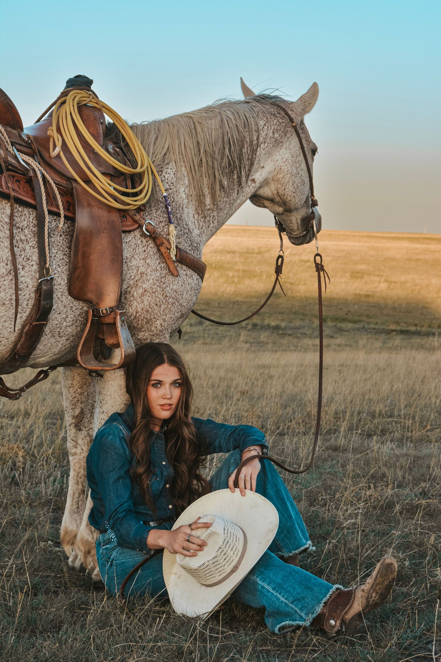 A dappled gray horse with western saddle stands near a seated cowgirl in denim at sunset on the prairie.