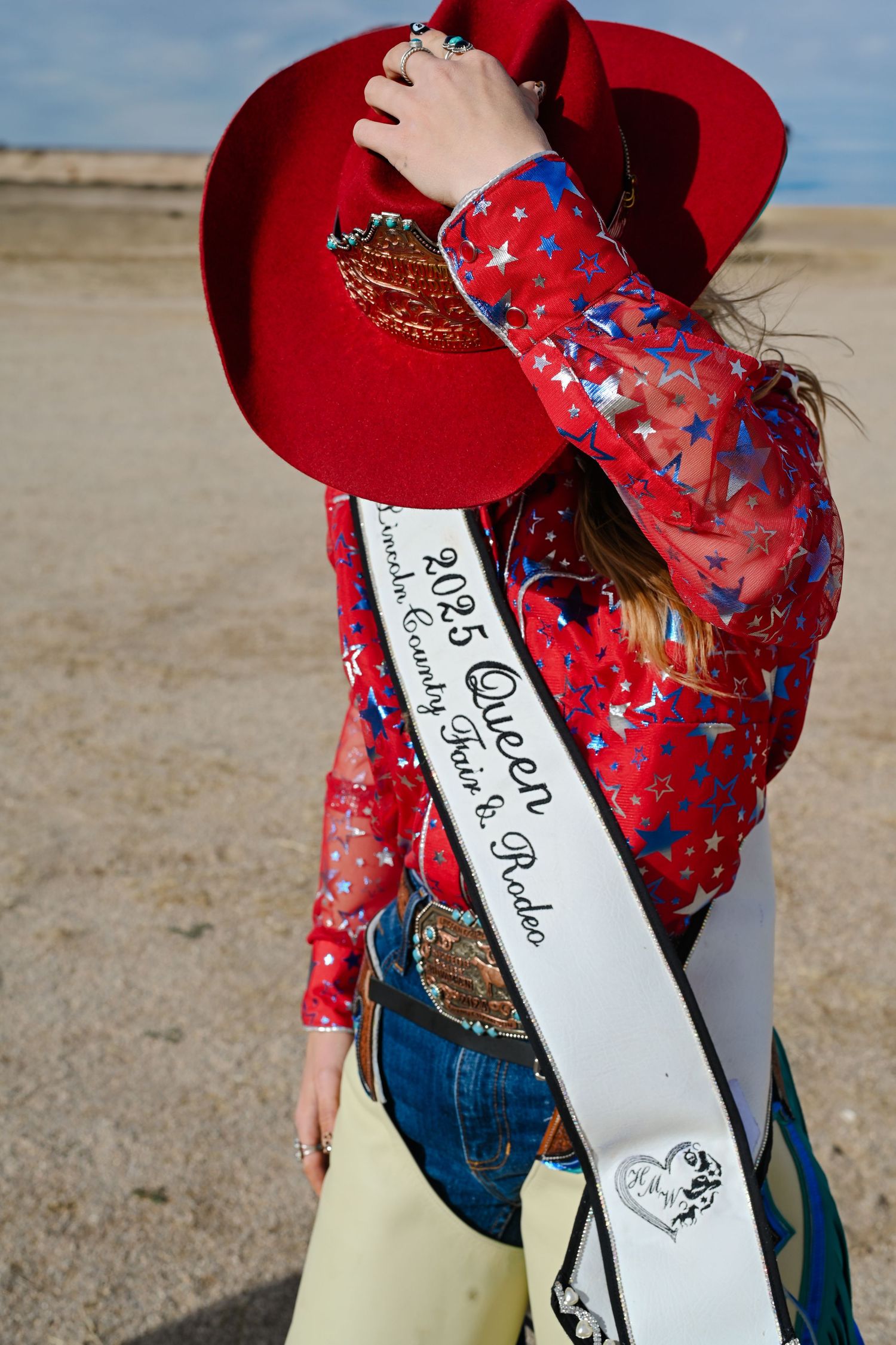 Western pageant contestant in red hat and floral blouse displays sash with awards at outdoor competition.