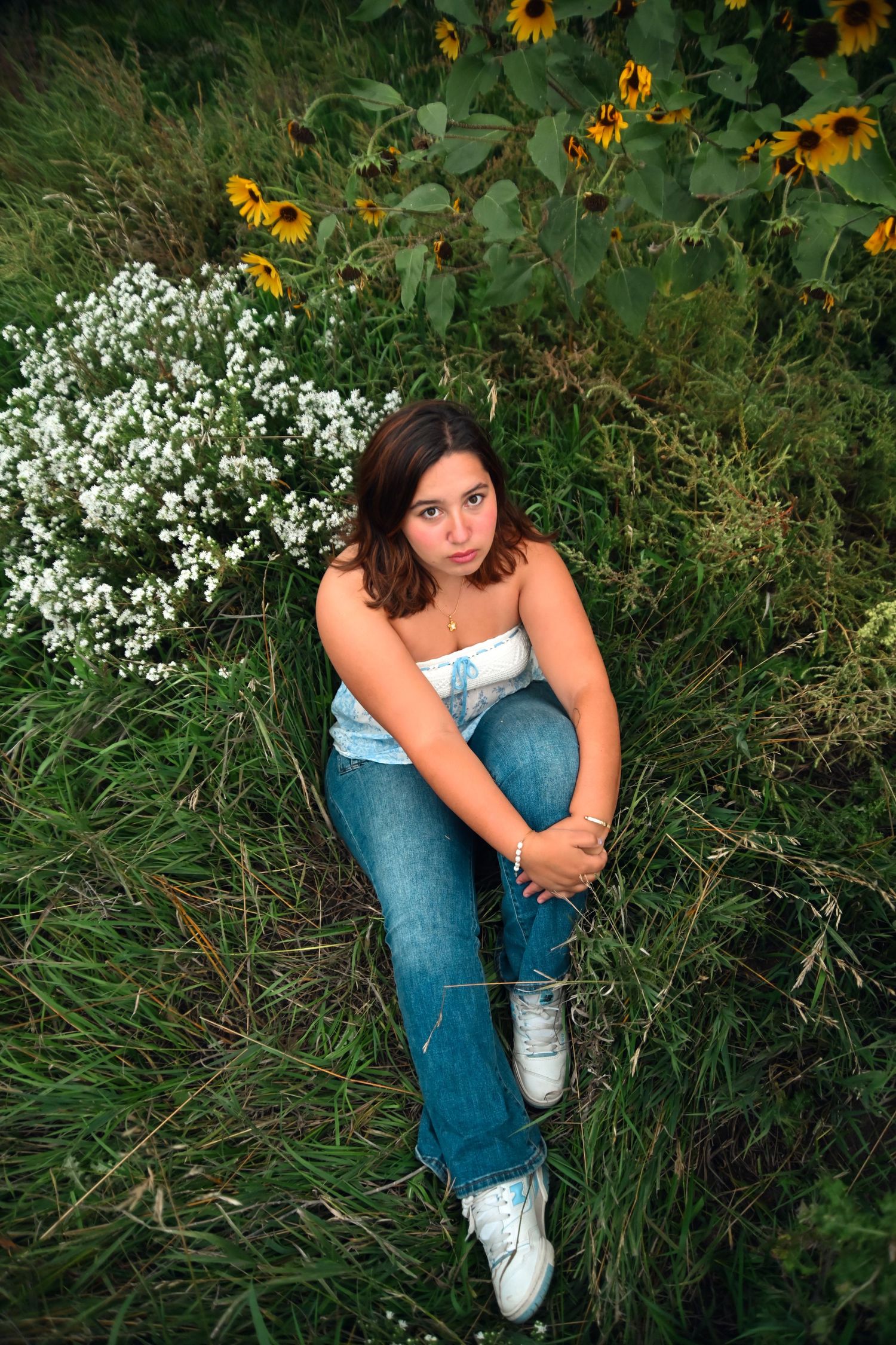 Young woman in jeans and white top sitting in a lush green field with sunflowers and white wildflowers.