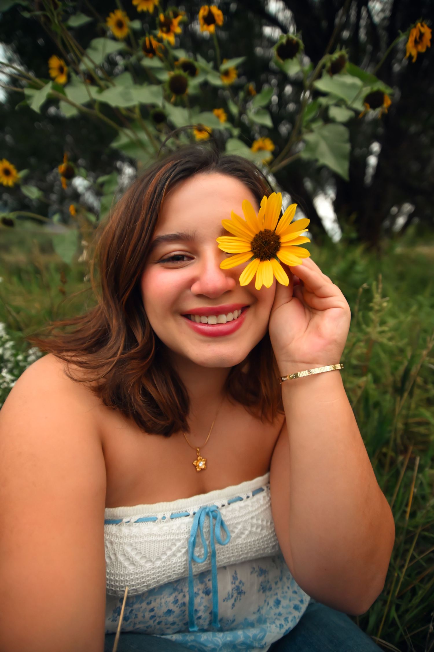 Smiling young woman holding a yellow sunflower over her eye outdoors near sunflower bushes.