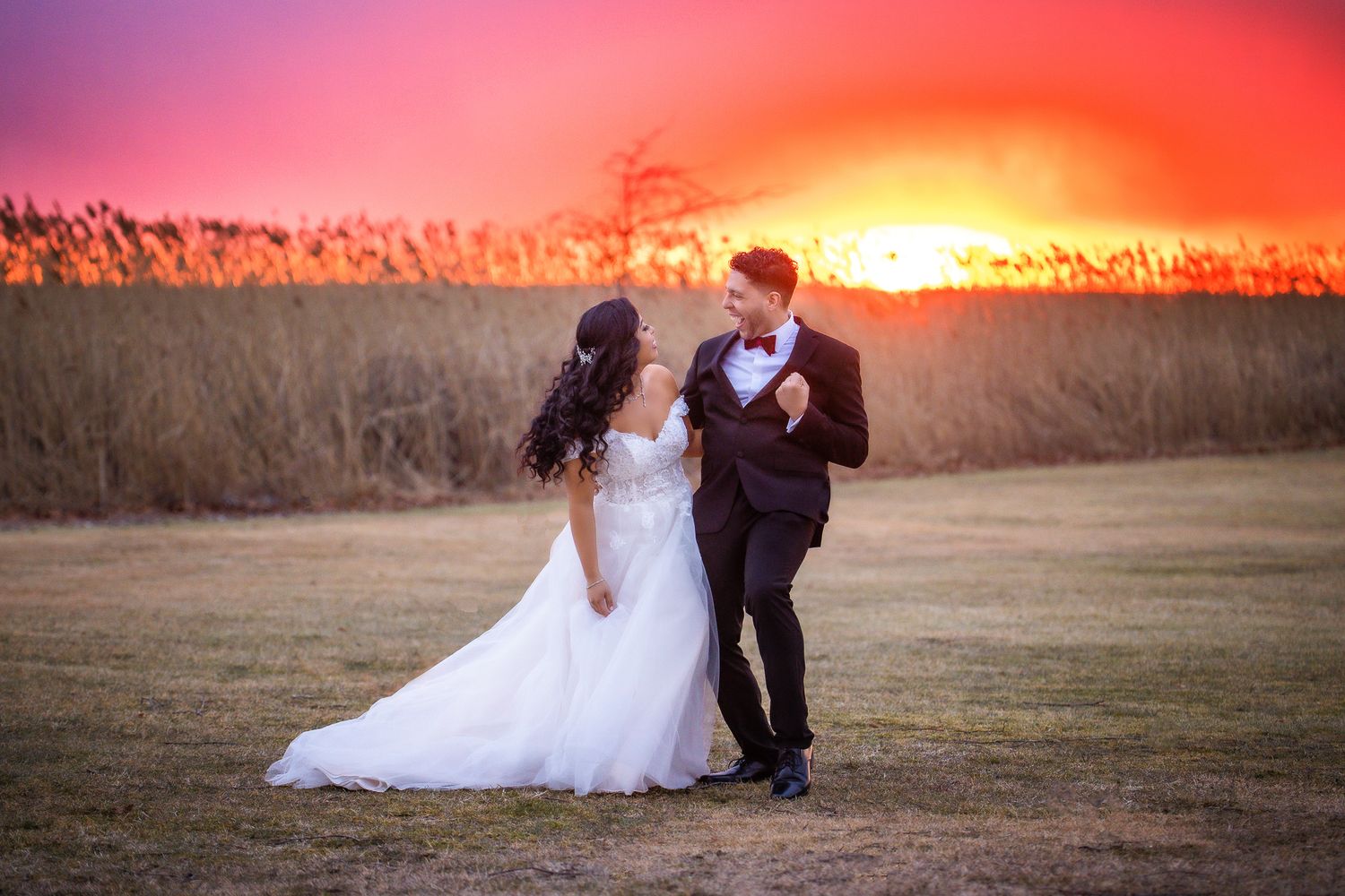 A romantic couple in wedding attire pose together in a field at sunset with dramatic orange sky and tall grass.