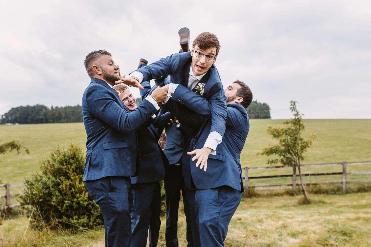 Groomsmen in blue suits playfully lift and support another member of the wedding party in an outdoor setting.