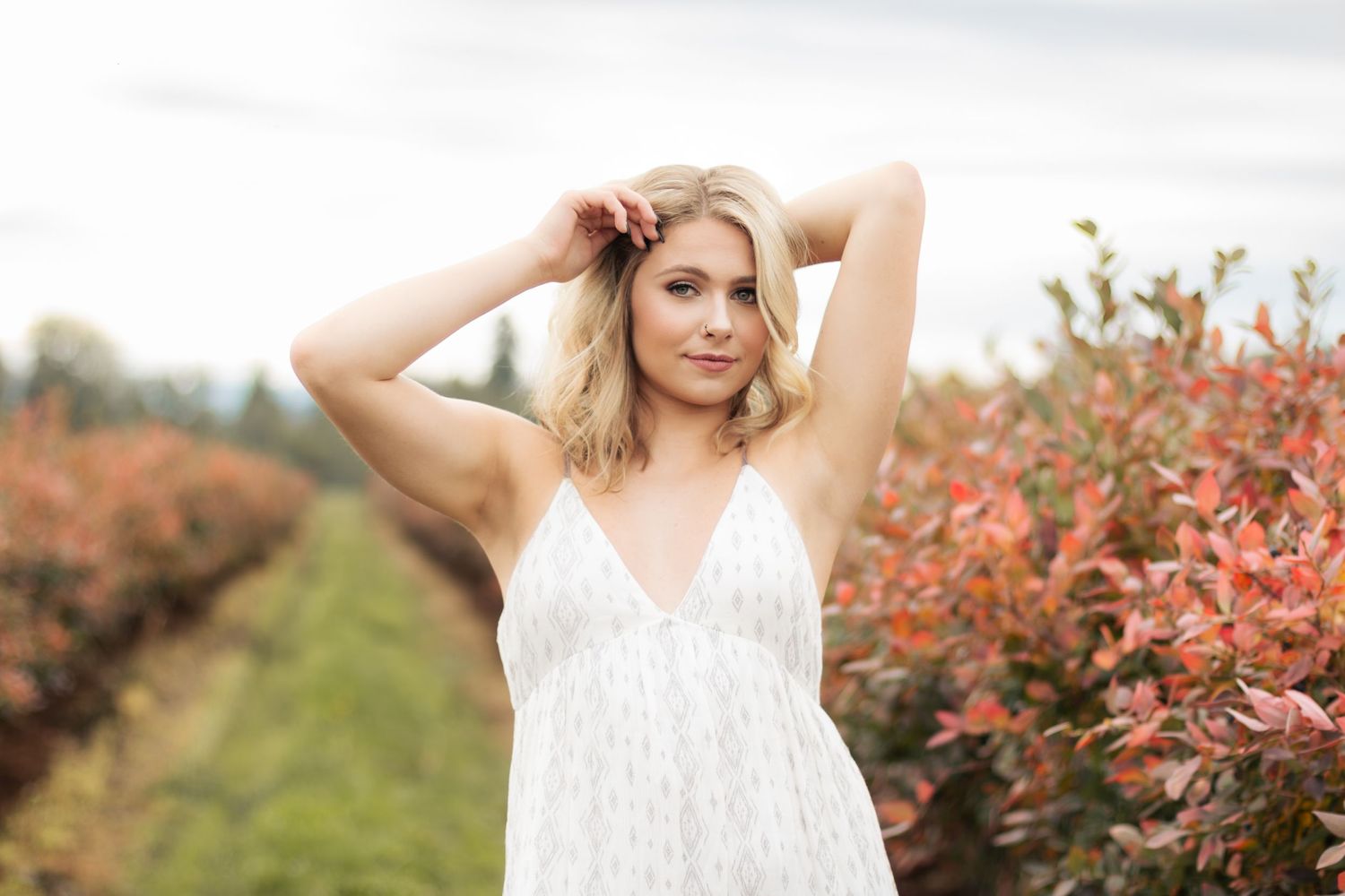 A woman in a white sundress poses outdoors near vibrant red autumn foliage in a garden setting.