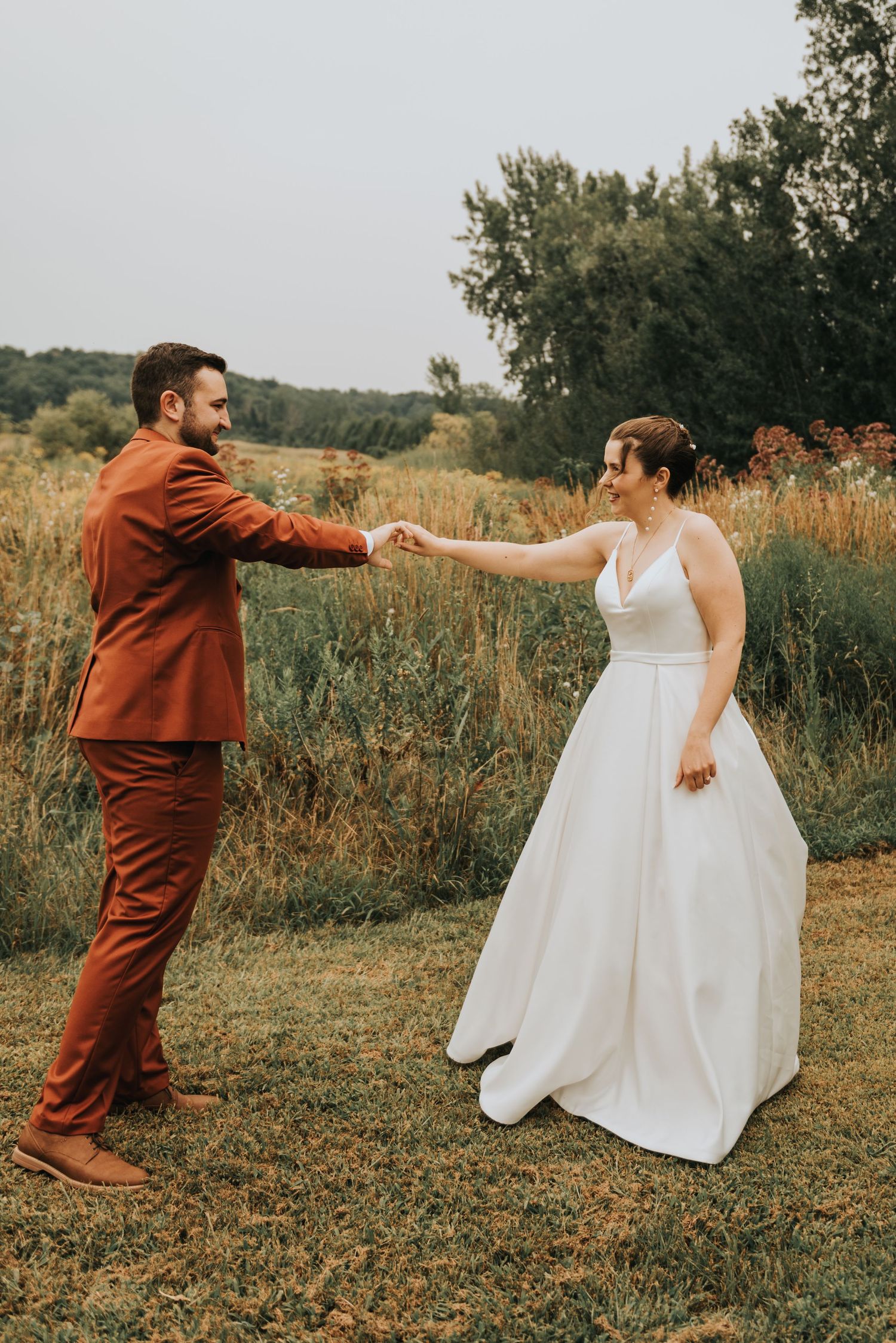 A couple dances together in a rustic outdoor setting, with the bride in white and groom in a rust-colored suit.