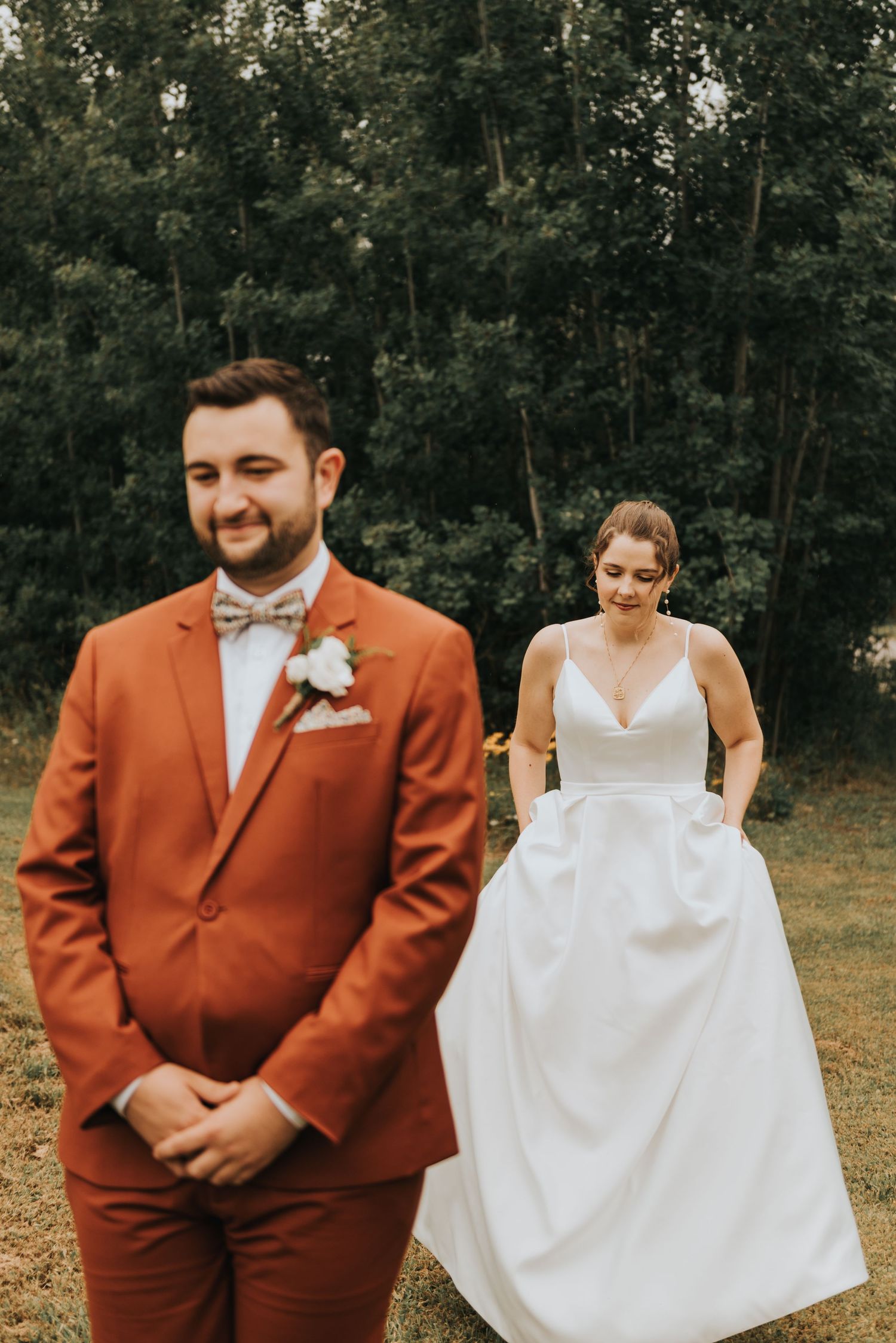 A wedding couple during their first look moment in a forest setting with the bride wearing white and groom in rust-colored suit.
