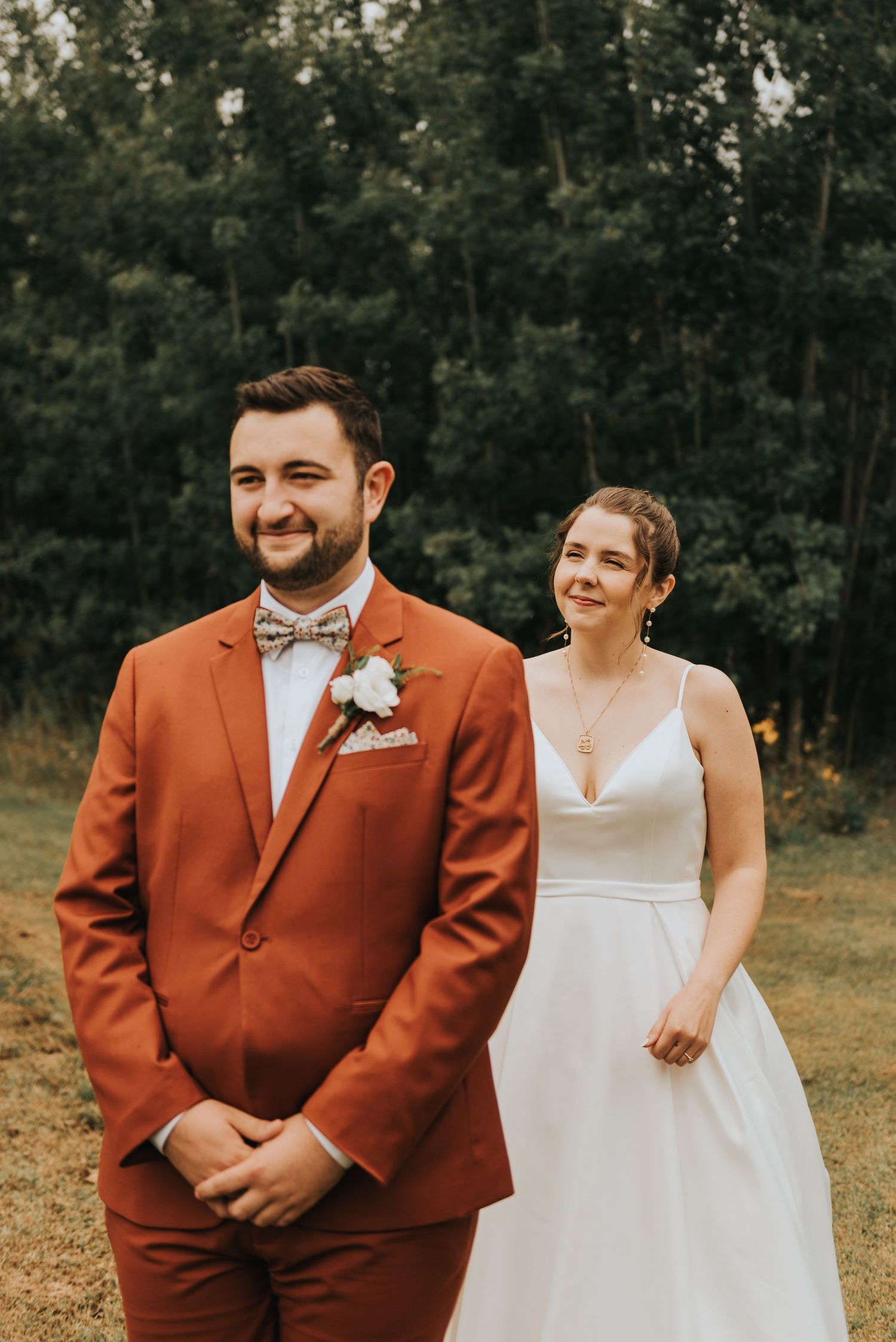 A couple smiles during a rustic outdoor wedding ceremony, with the groom wearing a burnt orange suit and bowtie.