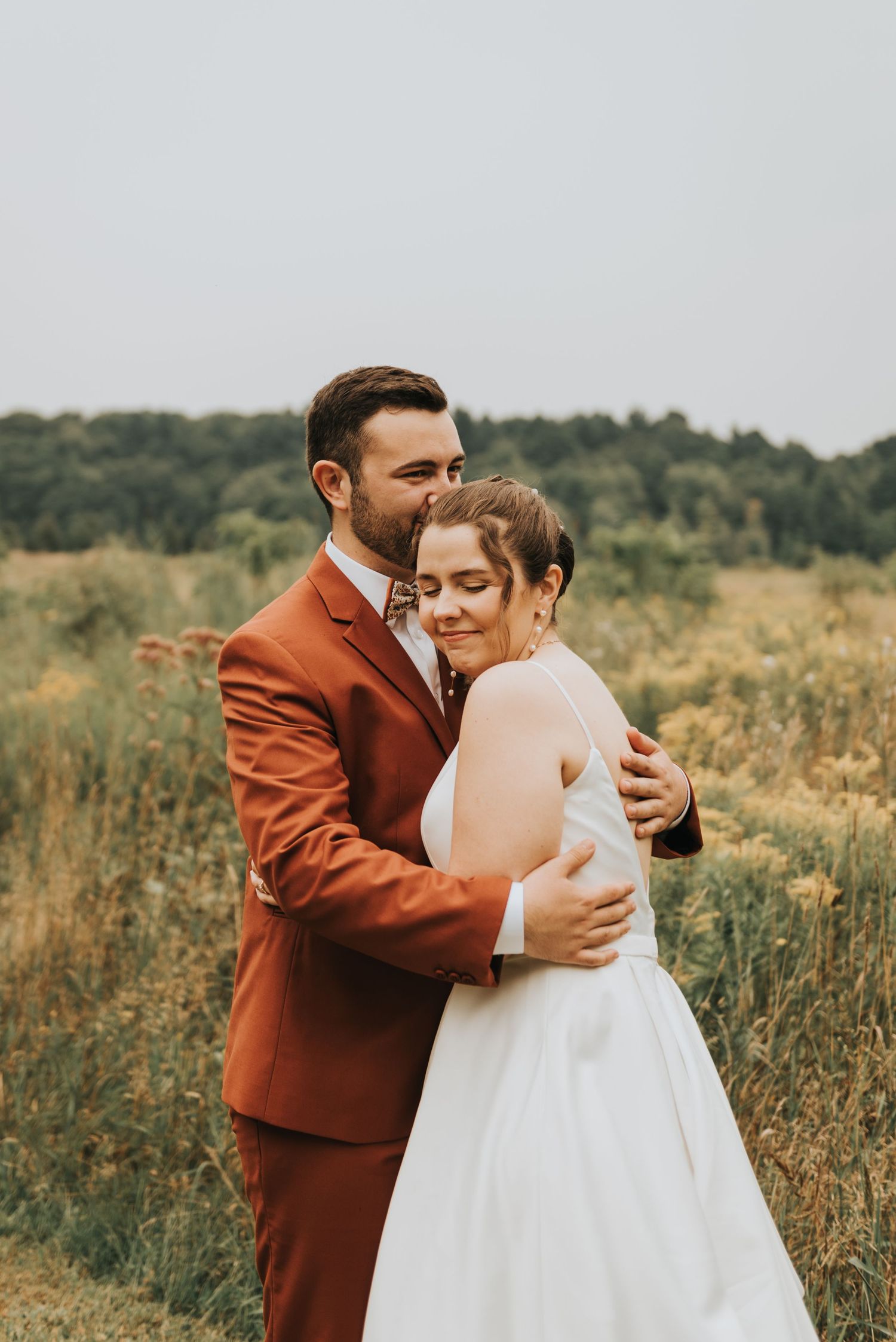 A couple in wedding attire embraces in a rustic field, the groom wearing a burnt orange suit and bride in white dress.