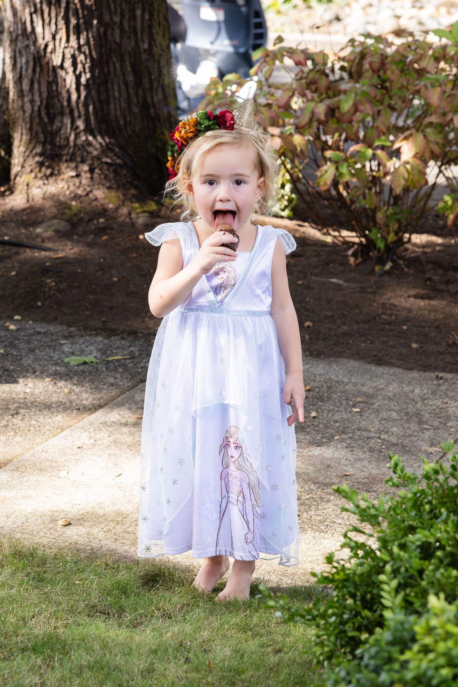A child in a white dress with a floral crown stands on a garden path while eating a snack outdoors.