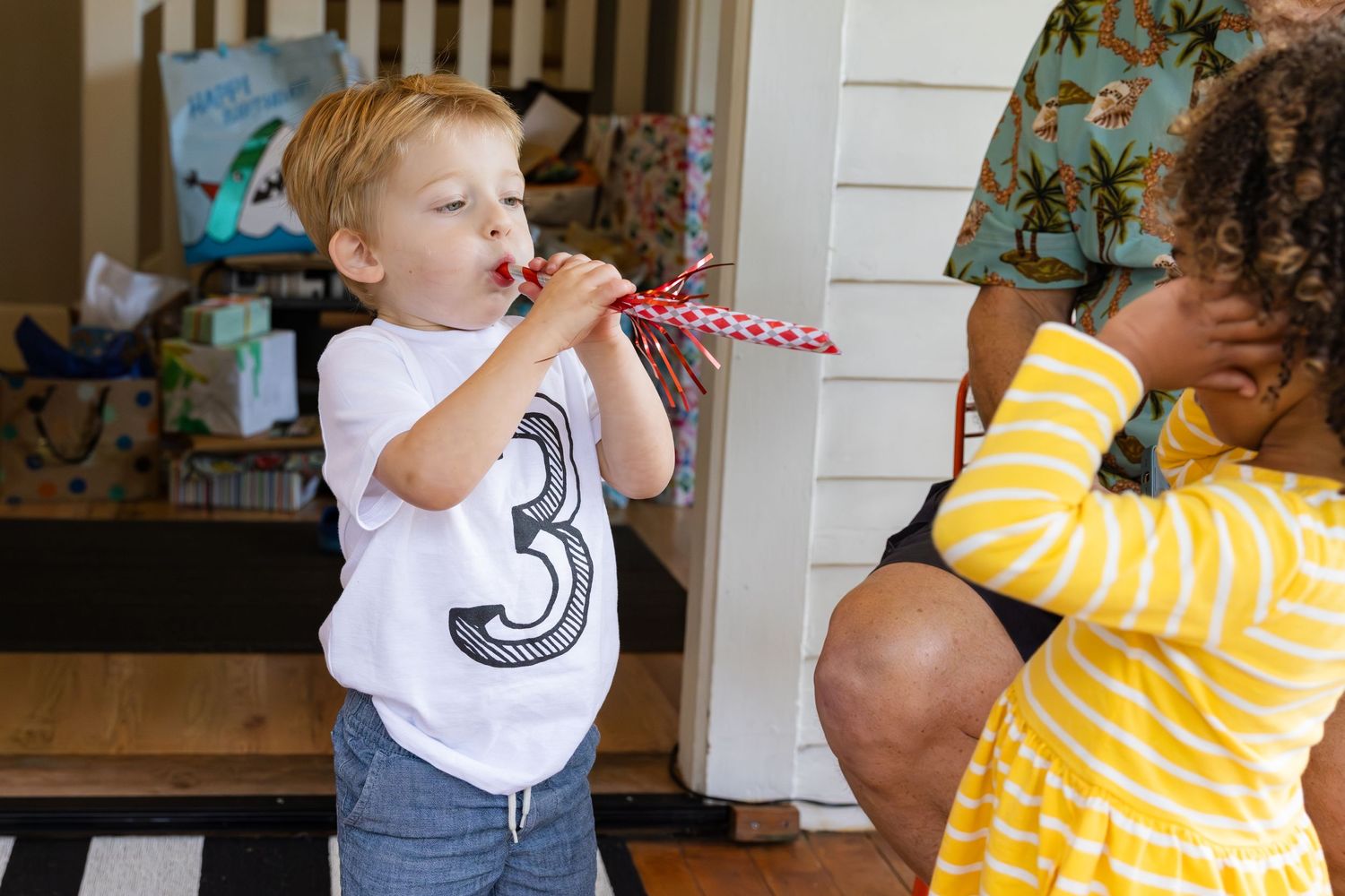Child in white shirt with number 3 plays a pink party horn while standing next to someone in yellow stripes.