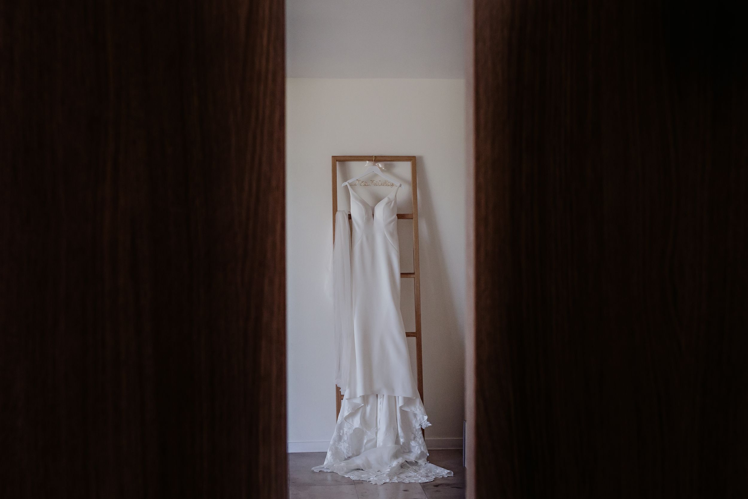 A white wedding dress hangs elegantly on a hanger against a plain white wall, viewed through a doorway.