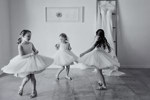 Three young children in white dresses twirling and dancing in bright room with wedding dress visible.