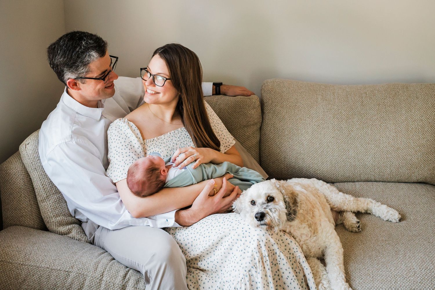 Family relaxes on gray couch with newborn baby and white fluffy dog in living room.