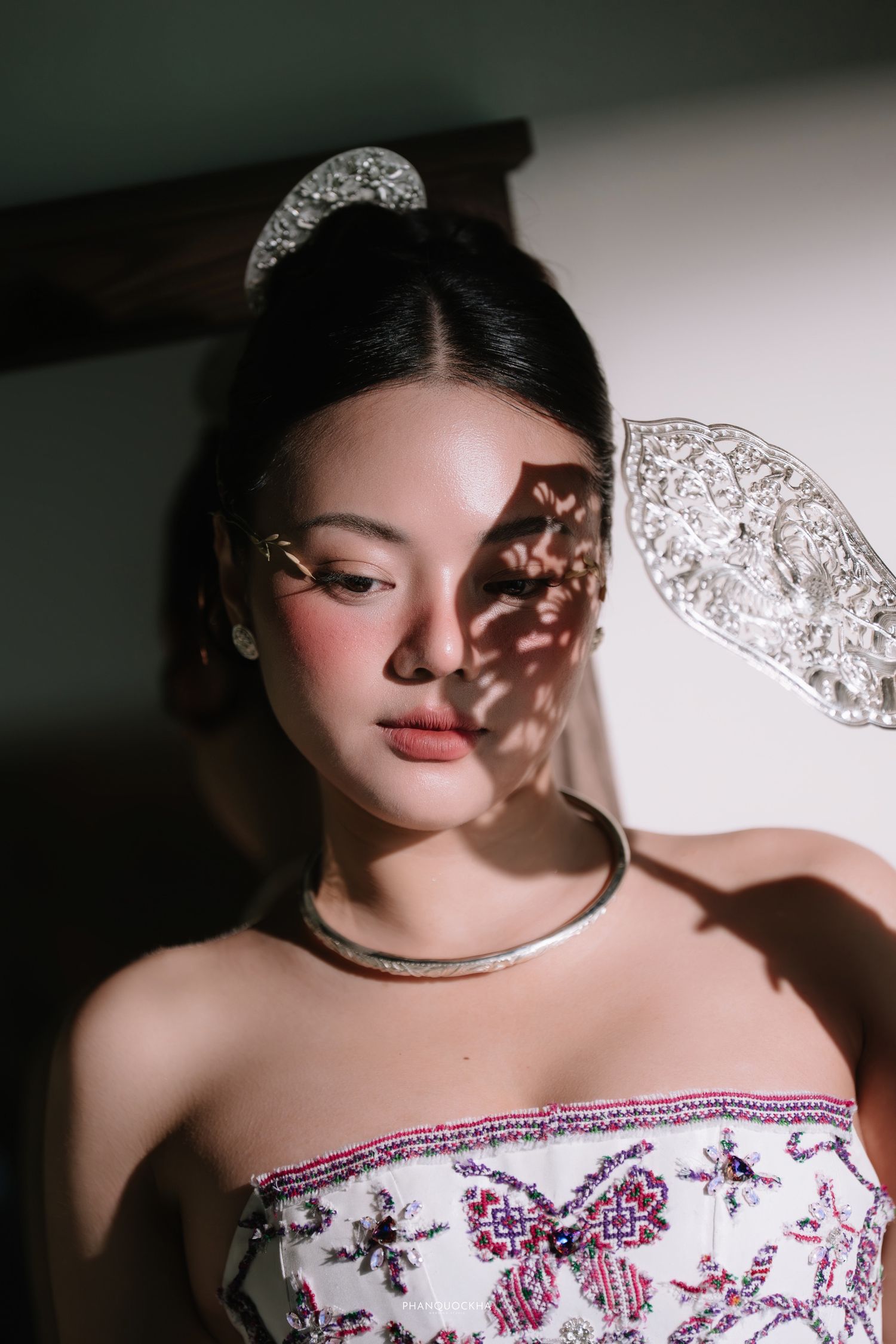 Beautiful Asian bride in embroidered strapless gown with crystal crown and lace fan accessory, bathed in warm natural light.
