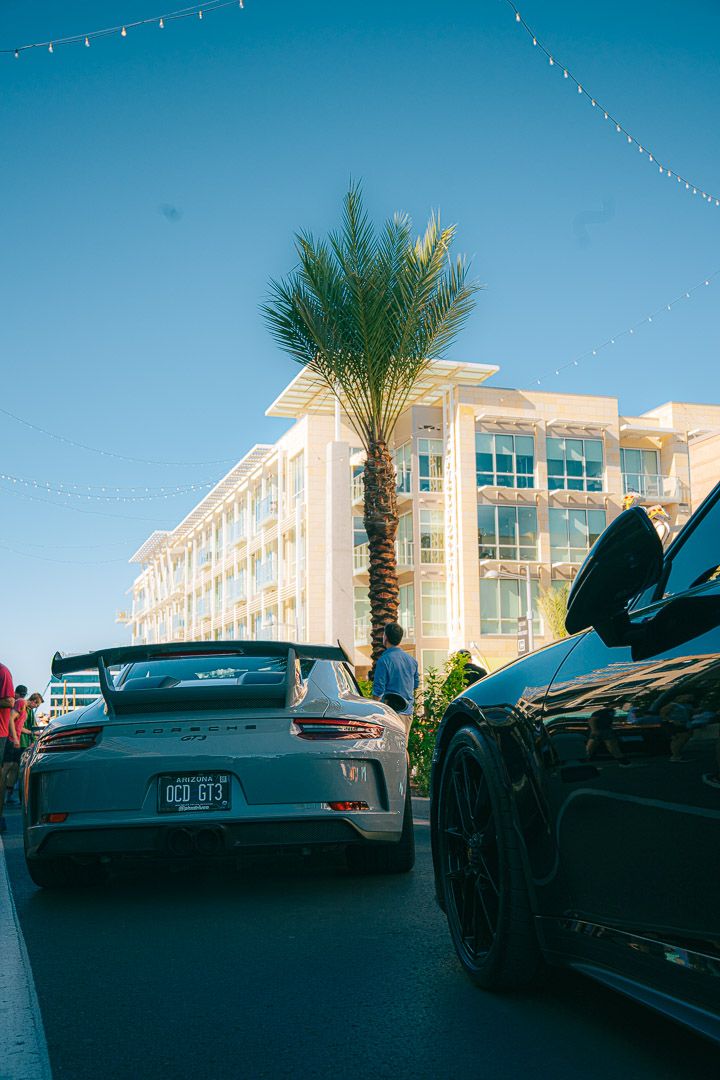 Luxury sports cars lined up at an outdoor car show with palm trees and string lights overhead.