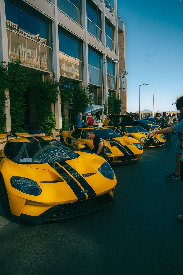 Row of bright yellow sports cars with black racing stripes parked outside commercial buildings.