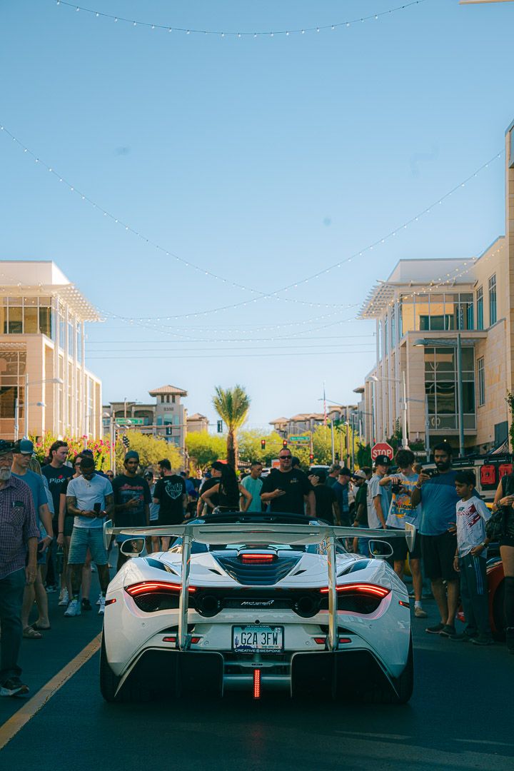 White McLaren supercar surrounded by spectators at a car show with coastal buildings in the background.