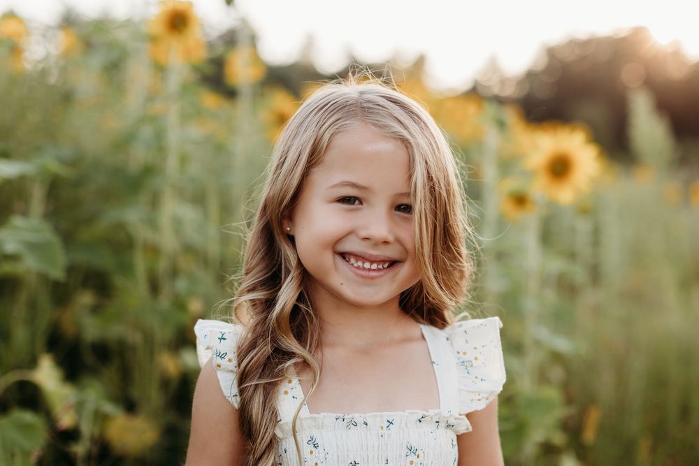 Child in white dress smiling brightly in a sunflower field during golden hour.