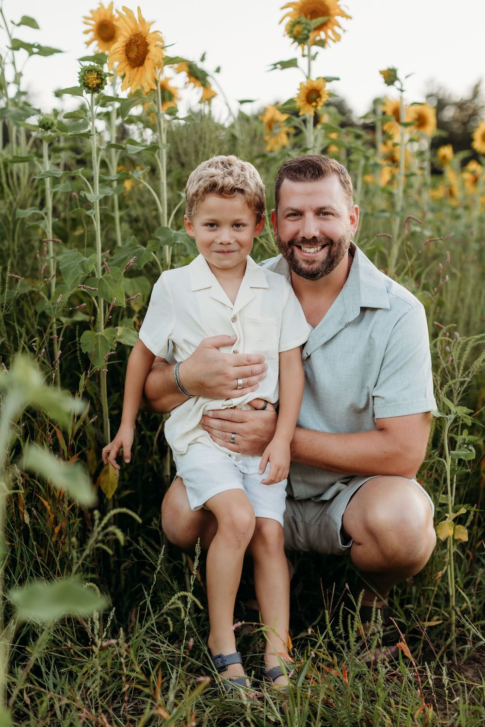Father and child sharing laughs while sitting in a sunflower field at sunset.